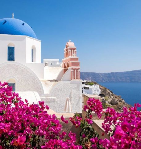 Red flowers and a whitewashed church with blue dome overlooking the sea in Santorini island.