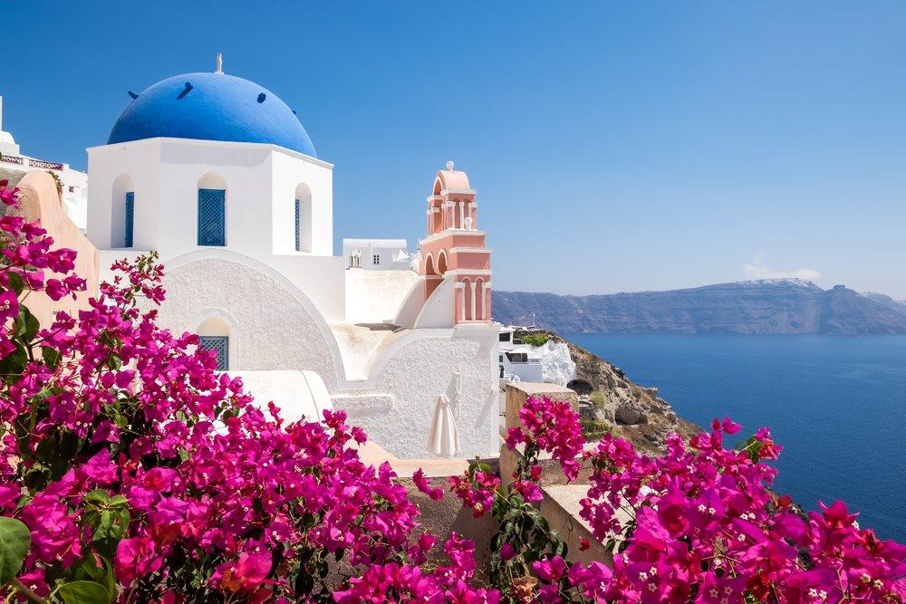 Red flowers and a whitewashed church with blue dome overlooking the sea in Santorini island.